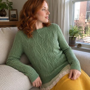 Può includere: Un maglione a maniche lunghe color verde salvia con un motivo a trecce verticali, scollo rotondo e polsini a coste. Il maglione è indossato sopra un indumento chiaro con un delicato bordo in pizzo sul fondo, abbinato a una gonna color senape.