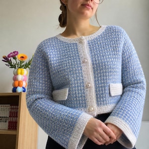 May include: A light blue and white textured knit cardigan featuring a round neckline, long sleeves with wide cuffs, two front pockets, and a front button closure with four round white pearl-like buttons. The garment has a consistent grid pattern.
