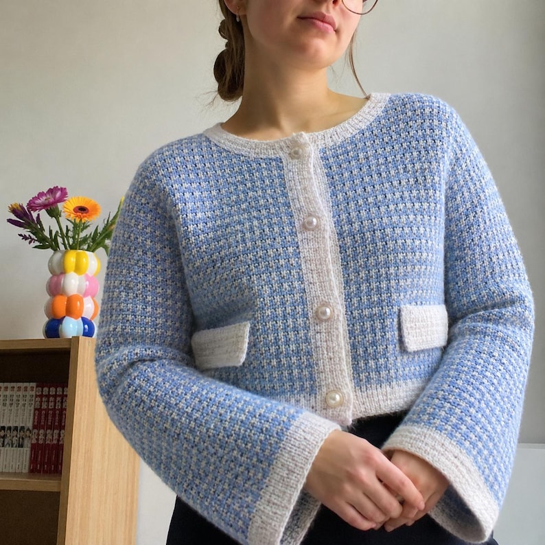 May include: A light blue and white textured knit cardigan featuring a round neckline, long sleeves with wide cuffs, two front pockets, and a front button closure with four round white pearl-like buttons. The garment has a consistent grid pattern.