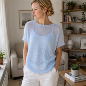 Könnte beinhalten: Ein hellblauer, kurzärmeliger Strickpullover mit einem zarten Lochmuster auf der Vorderseite und einem Rundhalsausschnitt. Das Kleidungsstück wird über einem passenden hellblauen Unterhemd getragen, was einen Lagen-Look mit weicher Textur erzeugt.