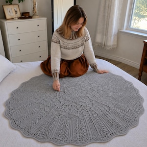 Könnte beinhalten: Eine große, kreisförmige, hellgraue Strickdecke mit einem komplizierten Spitzenmuster und einem gewellten Rand. Das Design umfasst ein zentrales sternförmiges Motiv, das sich in detaillierten, strukturierten Maschen nach außen erstreckt.