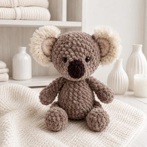May include: A handmade, plush koala bear in shades of brown and beige. The koala has a textured body, a dark brown nose, and fluffy ears. It is sitting on a cream-colored blanket, with white vases and a candle in the background.