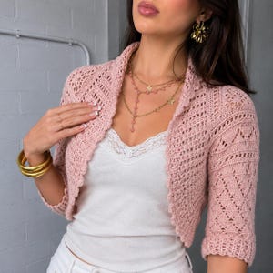 Può includere: Un bolero all'uncinetto rosa chiaro con maniche a tre quarti e apertura frontale, caratterizzato da un delicato motivo simile al pizzo su tutta la lavorazione. Il capo è indossato sopra una canotta bianca con bordo in pizzo sullo scollo.