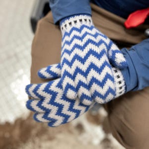 Könnte beinhalten: Ein Paar Strickhandschuhe mit einem sich wiederholenden Zickzackmuster in Blau und Cremeweiß. Die Handschuhe haben ein geripptes Bündchen am Handgelenk mit einem kleinen, dunklen geometrischen Detail am Rand für einen festen Sitz bei kaltem Wetter.