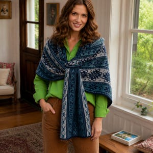 May include: A knitted shawl featuring a dark blue, light blue, and white geometric pattern with snowflake motifs. The shawl is draped over a bright green long-sleeved shirt and brown trousers. The fabric displays a dense, intricate jacquard knit.
