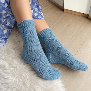 May include: A pair of mid-calf length knitted socks in a medium blue hue. The socks feature a repeating geometric lace pattern with vertical eyelet columns and a ribbed cuff at the top. The knit texture is consistent throughout the foot and ankle.
