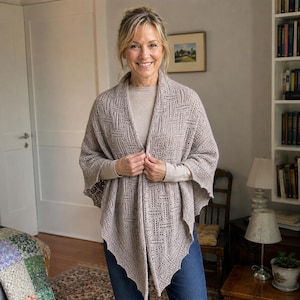 May include: A light beige knit shawl featuring an intricate geometric openwork pattern with a pointed hemline. The garment is worn over a matching beige long-sleeved top and blue denim jeans, showcasing a textured, lightweight construction.