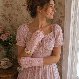 May include: A light pink knit dress with short puff sleeves and a square neckline, paired with matching light pink fingerless gloves featuring a delicate openwork lace pattern. The gloves are worn on the forearms, and a multi-strand pearl bracelet is visible.