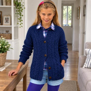 May include: A navy blue cable-knit cardigan featuring a V-neckline, long sleeves, and four large brown buttons down the front. The garment is worn over a blue and white plaid collared shirt and light blue denim shorts with purple leggings.