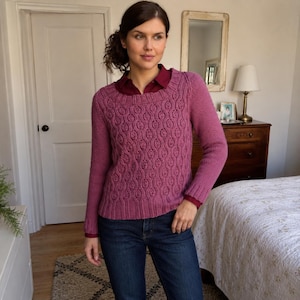 May include: A long-sleeved, mauve-coloured knit jumper featuring a textured, repeating oval pattern on the front panel, a ribbed crew neckline, and ribbed cuffs, worn over a dark red collared shirt and dark blue denim jeans.