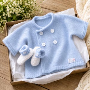 Peut inclure: Un gilet en tricot bleu clair à manches courtes avec quatre boutons blancs dans un style croisé, accompagné de deux chaussons en tricot blanc et bleu clair. Une petite étiquette rectangulaire blanche avec du texte et un cœur rouge est fixée.