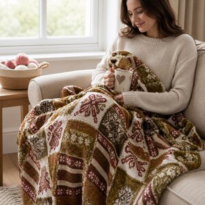 Può includere: Una coperta lavorata a maglia con un design patchwork a quadrati alternati, caratterizzata da intricati motivi a fiocchi di neve e disegni geometrici nei toni del verde oliva, marrone scuro e crema, appoggiata su un divano beige con una tazza.