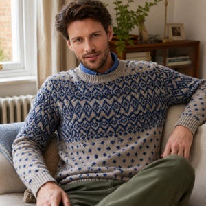 Pode incluir: Uma camisola bege de gola redonda com um padrão geométrico azul estilo fair isle no peito e ombros, com um padrão de cruzes azuis mais pequenas na parte inferior e nas mangas, usada sobre uma camisa de colarinho azul com punhos canelados.