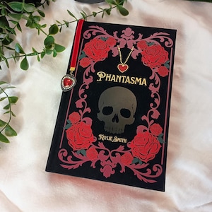 Puede incluir: Un libro negro titulado "Phantasma" con un diseño de calavera y rosas. La portada presenta rosas rojas y detalles dorados. Se adjunta un marcapáginas de terciopelo rojo con un dije en forma de corazón. Una vela encendida y vegetación en el fondo.