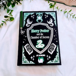May include: A hardcover book titled "Harry Potter and the Chamber of Secrets" by J.K. Rowling. The book has a black cover with green and white decorative elements, including a snake, stars, and phoenixes. A green bookmark with a spider charm is attached.