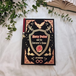 May include: A hardcover book titled "Harry Potter and the Sorcerer's Stone" by J.K. Rowling. The book has a black cover with gold and red accents, including an owl, wings, and a gemstone. A red bookmark with a charm is attached.