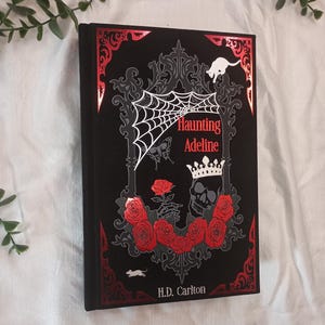 Könnte beinhalten: Ein schwarzes Hardcover-Buch mit dem Titel Haunting Adeline von H.D. Carlton. Der Einband zeigt rote Folienakzente, ein weißes Spinnennetz, einen Totenkopf mit Krone, rote Rosen, eine kleine weiße Katze und eine kleine weiße Maus.