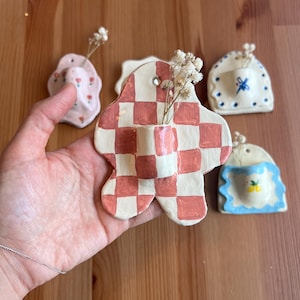 May include: A collection of handmade ceramic bud vases in various shapes and designs. One vase is held in a hand, featuring a red and white checkered pattern. Other vases include floral and geometric motifs, each holding dried flowers.