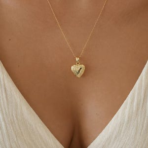 May include: A gold-colored heart-shaped pendant necklace. The pendant has a textured surface and hangs from a delicate chain. The necklace is worn against a cream-colored top.