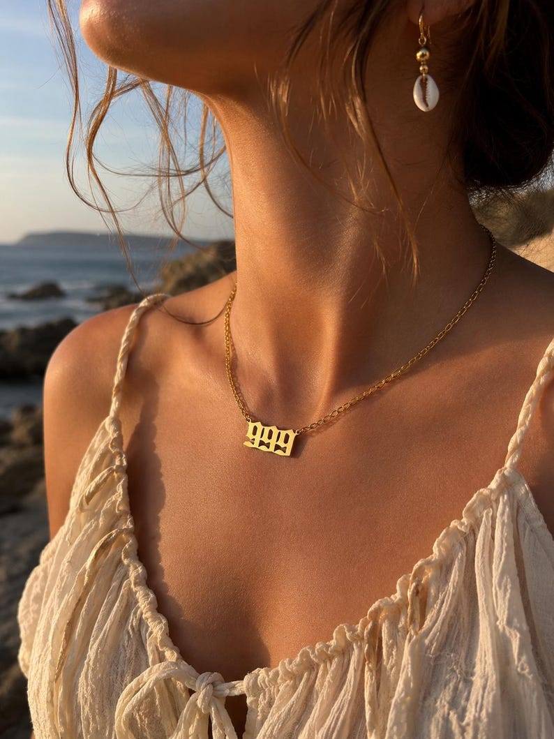以下が含まれることがあります： 数字「999」のペンダントが付いたゴールドカラーのネックレス。クリーム色のトップスの上から着用されています。貝殻のイヤリングが見えます。背景にはビーチと海が写っています。