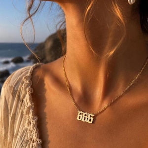 以下が含まれることがあります： ゴシック体の数字「666」が付いたゴールドカラーのネックレス。繊細なチェーンが付いています。背景には海とクリーム色のトップスを着た人の姿が映っています。