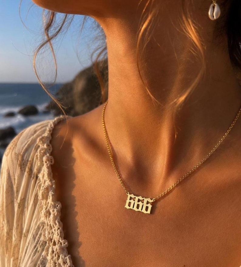 以下が含まれることがあります： ゴシック体の数字「666」が付いたゴールドカラーのネックレス。繊細なチェーンが付いています。背景には海とクリーム色のトップスを着た人の姿が映っています。