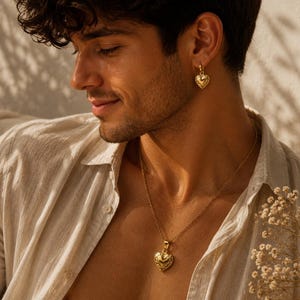 May include: A gold heart-shaped pendant necklace and matching earrings. The jewelry has a textured surface and is worn with a light-colored button-down shirt. The image is lit with warm sunlight.