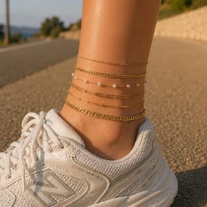 May include: A close-up of an ankle wearing several gold anklets. The anklets have different designs, including fine chains, pearls, and rectangular links. The person is wearing a white trainer.