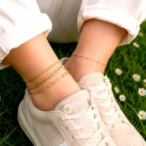 May include: Gold anklets in various chain styles adorn the ankles. The anklets are layered, with a delicate beaded chain and thicker curb and paperclip chains. The person is wearing white pants and beige sneakers, with a green grass background.