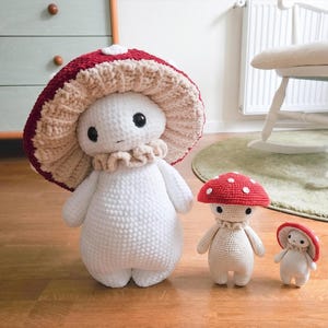 May include: Three crochet mushroom figures in varying sizes. The largest is white with a red and tan mushroom cap. The smaller figures have red caps with white spots and tan bodies. The figures have black eyes and tan ruffs.