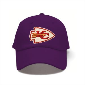 May include: A purple baseball cap featuring an embroidered arrowhead-shaped patch on the front. The patch is cream-colored with a red border and contains the red letters KC with a yellow arrow pointing to the right across the center of the letters.