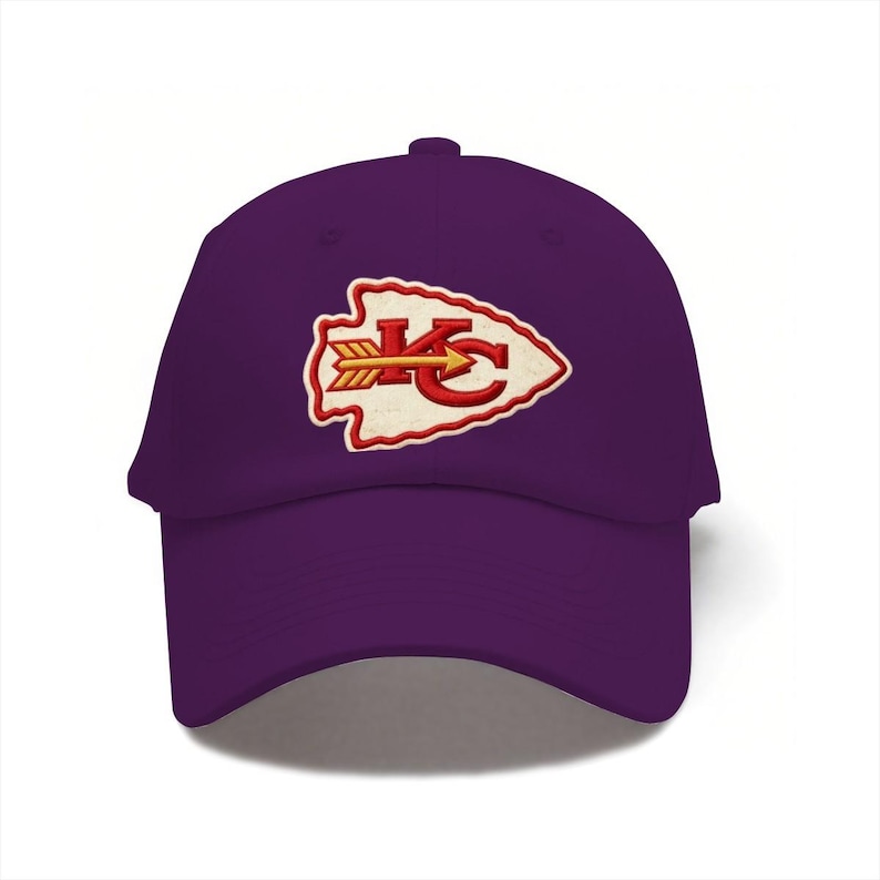May include: A purple baseball cap featuring an embroidered arrowhead-shaped patch on the front. The patch is cream-colored with a red border and contains the red letters KC with a yellow arrow pointing to the right across the center of the letters.