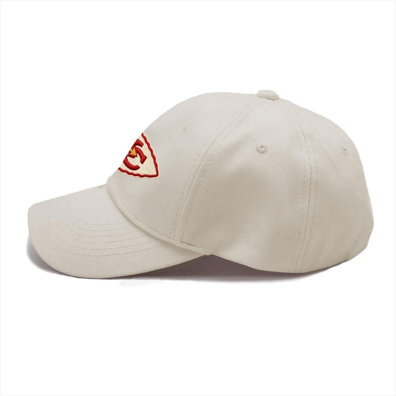 May include: A cream-colored baseball cap featuring an embroidered logo on the front panel. The logo consists of a red arrowhead shape with a yellow border and the red letters KC centered inside. The cap has a curved brim and stitched panels.