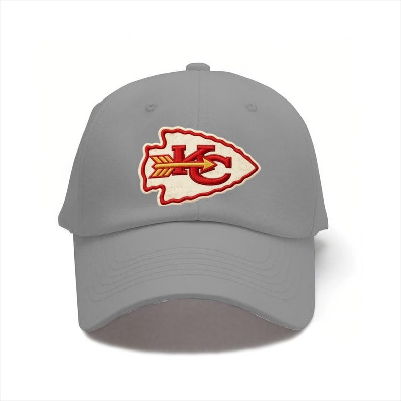 May include: A light gray baseball cap featuring an embroidered patch on the front. The patch is shaped like an arrowhead with a white background, a red border, and the red letters KC with a yellow and red arrow pointing horizontally across them.