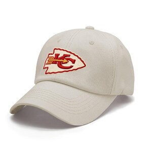 May include: A cream-colored baseball cap featuring an embroidered arrowhead-shaped patch on the front. The patch displays the red letters KC with a yellow and red arrow running horizontally through the center of the design on a white background.