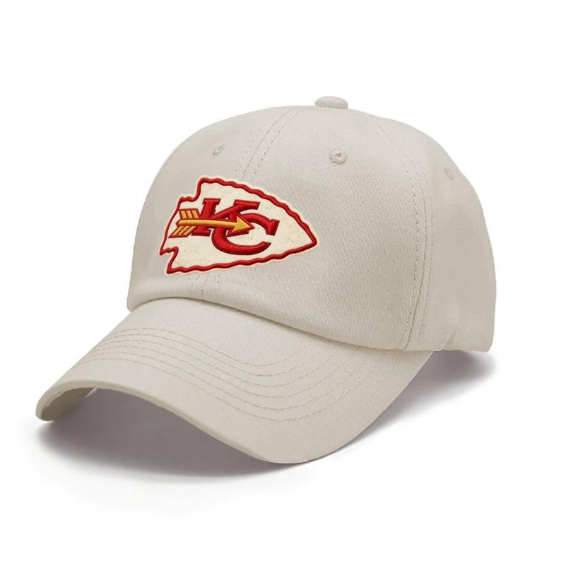 May include: A cream-colored baseball cap featuring an embroidered arrowhead-shaped patch on the front. The patch displays the red letters KC with a yellow and red arrow running horizontally through the center of the design on a white background.