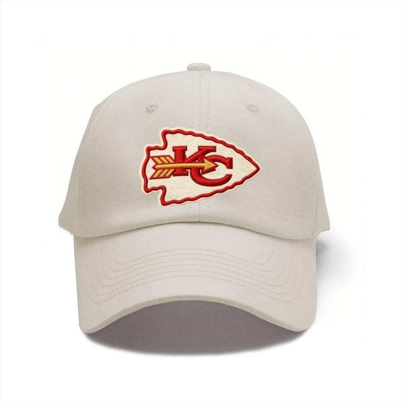 May include: A cream-colored baseball cap featuring an embroidered patch on the front. The patch is shaped like an arrowhead with a red border, containing the red letters KC and a yellow arrow pointing to the right across the center of the letters.
