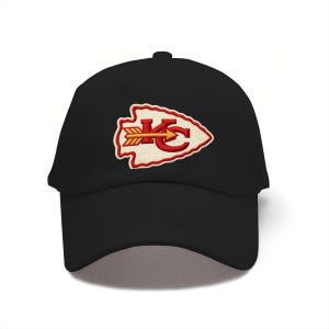 May include: A black baseball cap featuring an embroidered patch on the front. The patch is shaped like an arrowhead with a cream-colored center, a red border, and the red letters KC with a yellow arrow pointing horizontally through the letters.