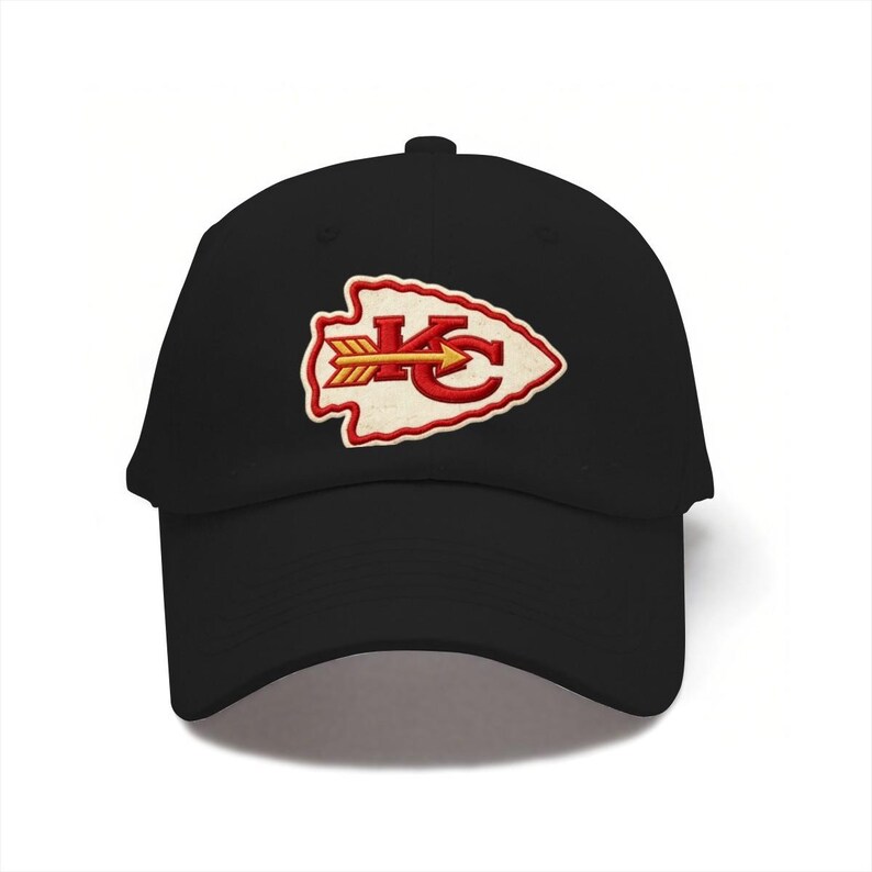 May include: A black baseball cap featuring an embroidered patch on the front. The patch is shaped like an arrowhead with a cream-colored center, a red border, and the red letters KC with a yellow arrow pointing horizontally through the letters.