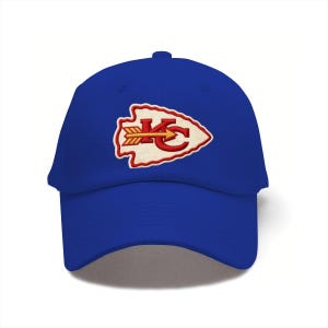 May include: A royal blue baseball cap featuring an embroidered patch on the front. The patch is shaped like an arrowhead with a cream-colored background, a red border, and the red letters KC with a yellow and red arrow pointing to the right.