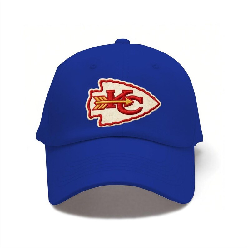 May include: A royal blue baseball cap featuring an embroidered patch on the front. The patch is shaped like an arrowhead with a cream-colored background, a red border, and the red letters KC with a yellow and red arrow pointing to the right.