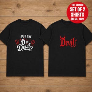 Könnte beinhalten: Zwei schwarze T-Shirts mit kurzen Ärmeln. Das linke Shirt zeigt den weißen Schriftzug I PUT THE D IN Devil mit roten Dreizack-Symbolen. Das rechte Shirt zeigt den roten Schriftzug Devil mit kleinen roten Hörnern über dem D und einem Schwanz.