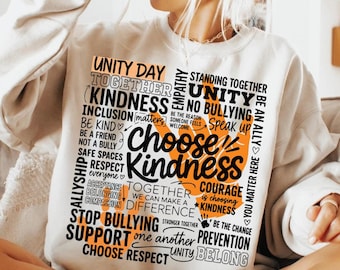 Puede incluir: Una sudadera beige de manga larga con un gran estampado gráfico en negro y naranja. El diseño consta de varias palabras y frases, incluyendo "Choose Kindness", "Unity Day", "No Bullying" y "Together We Can Make A Difference".