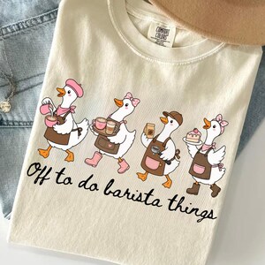 Puede incluir: Una camiseta de color crema con un gráfico de cuatro gansos blancos que llevan delantales y accesorios, cargando artículos relacionados con el café como tazas, un portafiltros y un trozo de pastel. El texto debajo dice, Off to do barista things.