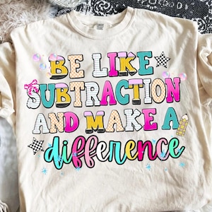 May include: A cream-coloured short-sleeve t-shirt featuring the colourful text BE LIKE SUBTRACTION AND MAKE A difference. The graphic includes pencil-shaped letters, checkered stars, small pink bows, and blue sparkles on the front of the shirt.