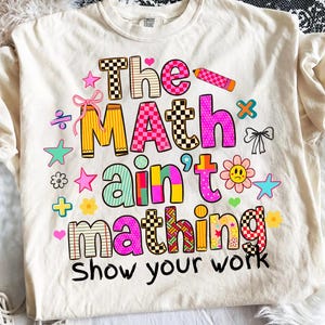 May include: A cream-coloured t-shirt featuring a colourful graphic with the text "The MAth ain't mathing Show your work". The design includes various patterns like checkers, stripes, and polka dots in pink, yellow, blue, and orange, alongside small icons of stars, bows, flowers, and a pencil.