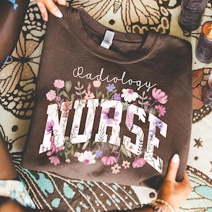 Pode incluir: Um moletom marrom com as palavras "Radiology Nurse" em letras brancas, cercado por um design floral em rosa, roxo e branco. O moletom está sendo segurado por uma pessoa.