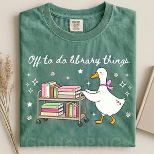 Puede incluir: Una camiseta verde salvia con el gráfico de una oca blanca con un lazo rosa, empujando un carrito de metal lleno de libros coloridos. El texto "Off to do library things" aparece impreso encima, rodeado de pequeñas estrellas y flores blancas.