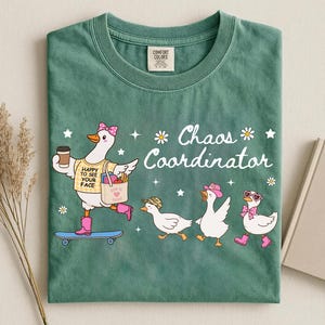 May include: A sage green t-shirt featuring a graphic of a duck on a skateboard wearing a yellow shirt that says HAPPY TO SEE YOUR FACE, holding a coffee and a tote bag. Three smaller ducks follow, with the text Chaos Coordinator and white daisies.