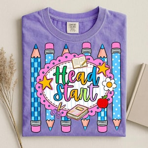 May include: A purple t-shirt featuring a graphic print with the text Head Start in colorful letters. The design includes blue pencils, a pink scalloped border, an open book, a closed book, a pair of scissors, a yellow star, a flower, and an apple.